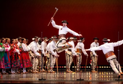Na pierwszym planie skaczący Artysta trzymający w ręku ciupagę, w tle Artyści w kostiumach podhalańskich podczas wykonywania choreografii tanecznej
