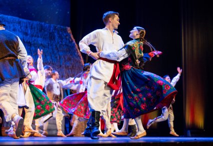 Zbliżenie na parę Artystów w kostiumach ludowych podczas wykonywania choreografii tanecznej w tle reszta część Artystów