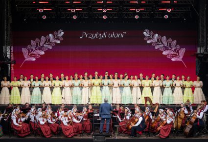 Widok na scenę na której znajdują się Artyści Chóru w kostiumach stylowych z końca XIX wieku wraz z Orkiestrą w kostiumach wilanowskich i Dyrygentem trzymającym w ręku batutę