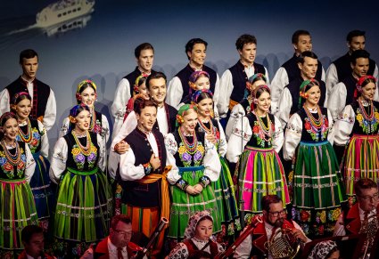 Zbliżenie na Artystów z chóru w kostiumach łowickich ustawionych w rzędach, na dole zdjęcia widoczna część orkiestry