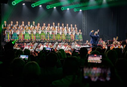 Widok na scenę na której znajdują się Artyści Chóru w kostiumach łowickich, niżej Artyści Orkiestry w kostiumach wilanowskich oraz Dyrygent zwrócony w ich stronę