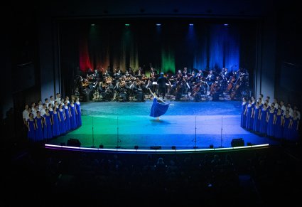 Na środku sceny Artystka Tancerka w niebieskiej sukience w trakcie wykonywania choreografii tanecznej. Po bokach sceny Artyści Chóru. Na tyłach sceny Orkiestra wraz z Dyrygentem