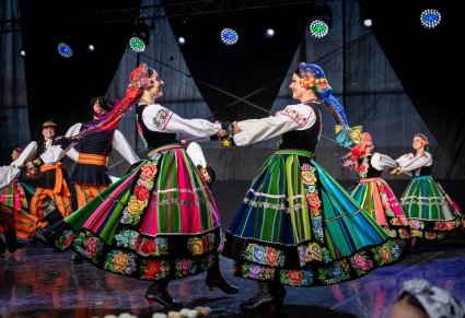 Na pierwszym planie dwie Artystki w kostiumach łowickich trzymające się za ręce w trakcie wykonywania choreografii tanecznej