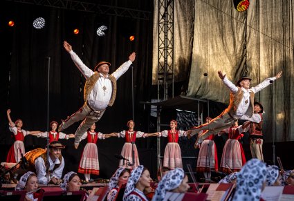 Dwóch Artystów wykonujących spektakularną akrobacje taneczną a w tle artystki trzymające się za ręce. Artyści ubrani są w kostiumy jurgowskie