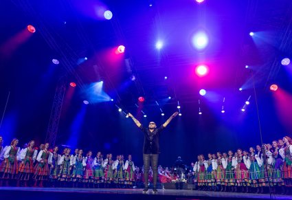 Na środku scena wokalista zespołu Kombii, po jego bokach Artyści zespołu "Mazowsze" w kostiumach ludowych, w tle widoczna część orkiestry wraz z Dyrygentem