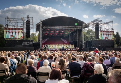Widok na Publiczność i na scenę plenerową na której znajdują się Artyści Chóru w kostiumach stylowych z końca XIX wieku wraz z Orkiestrą w kostiumach wilanowskich i Dyrygentem trzymającym w ręku batutę