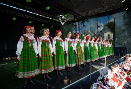 Artyści na scenie w kostiumach kurpiowskich ustawieni w rzędzie a na dole widoczna część orkiestry