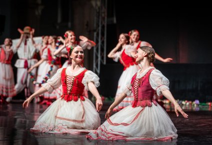Dwie Artystki na pierwszym planie wykonujące choreografię taneczną na scenie w kostiumach stylizowanych na kostium krakowski w tle reszta Artystek również w tych samych kostiumach