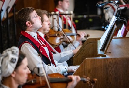 Zbliżenie na akompaniatora w kostiumie wilanowskim, widoczni również pozostali Artyści Orkiestry grający na instrumentach smyczkowych