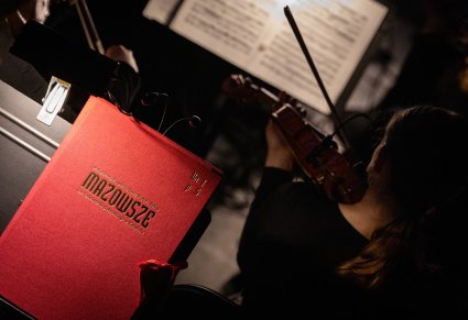 Zbliżenie na Muzyczkę grającą na instrumencie smyczkowym, widoczna również czerwona teczka na nuty ze złotym napisem nazwy zespołu