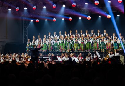 Na pierwszym planie Dyrygent, na scenie Artyści Chóru w kostiumach łowickich, niżej Artyści Orkiestry w kostiumach wilanowskich
