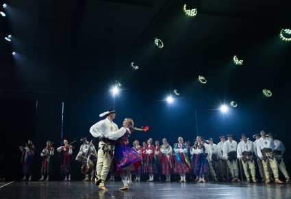 Na pierwszym planie para Artystów w kostiumach podhalańskich w trakcie wykonywania choreografii tanecznej.