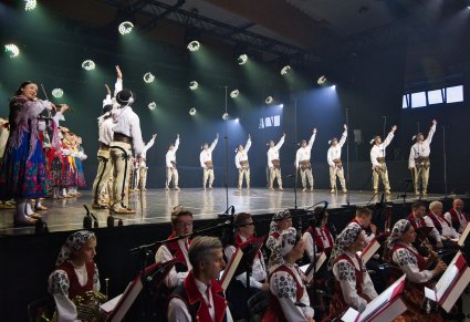 Na scenie Artyści w kostiumach podhalańskich w trakcie wykonywania choreografii tanecznej, niżej Artyści Orkiestry w kostiumach wilanowskich