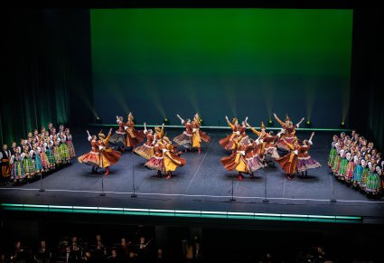 Na scenie Artyści w kontuszach w trakcie wykonywania choreografii tanecznej, po bokach sceny Artyści chóru w kostiumach łowickich podczas śpiewu