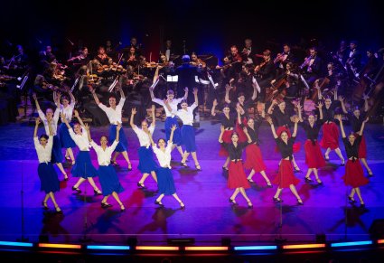 Na scenie Artystki Tancerki w trakcie wykonywania choreografii tanecznej, ubrane w kostiumy współczesne, w tle Orkiestra