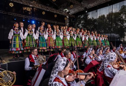 Na scenie Artyści Chóru w kostiumach łowickich ustawieni w dwurzędzie, niżej sceny widoczna część Artystów Orkiestry w kostiumach wilanowskich, grających na instrumentach