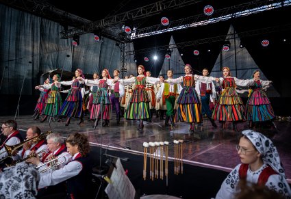 Grupa Artystów na scenie w kostiumach opoczyńskich wykonująca choreografię taneczną . Widoczna część orkiestry pod sceną