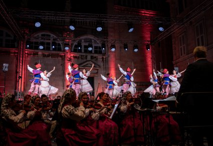 Na pierwszym planie Orkiestra a w tle Artyści w kostiumach rondo na scenie w trakcie wykonywania choreografii tanecznej