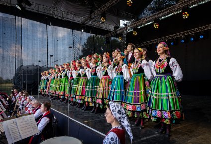 Na scenie Artyści Chóru w kostiumach łowickich, niżej sceny widoczna część Artystów Orkiestry w kostiumach wilanowskich grająca na instrumentach