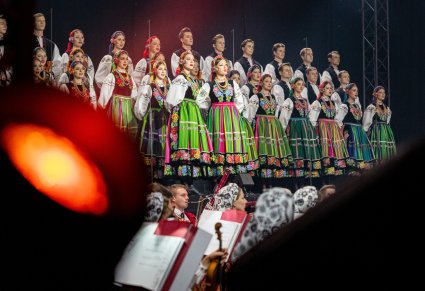 Na pierwszym planie lampa od scenografii, w tle Artyści chóru w kostiumach łowickich