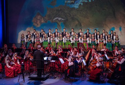 Na pierwszym planie Dyrektor Jacek Boniecki zwrócony w stronę orkiestry. Za orkiestrą Artyści chóru w kostiumach łowickich ustawieni w paru rzędach