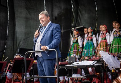 Na pierwszym planie Dyrektor Jacek Boniecki wygłaszający mowę do mikrofonu, a w tle Artyści z chóru w kostiumach łowickich