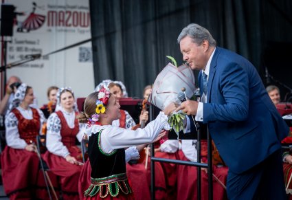 Na pierwszym planie mała dziewczynka ubrana w kostium regionalny wręcza kwiaty Dyrektorowi Jackowi Bonieckiemu w tle Orkiestra