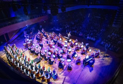 Widok z góry na scenę, a na niej Artyści Chóru w kostiumach łowickich, Orkiestra w kostiumach wilanowskich i Dyrygent z batutą. Widoczna część publiczności