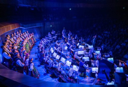 Boczny widok na scenę a na niej Artyści Chóru w kostiumach łowickich, Orkiestra wraz z Dyrygentem