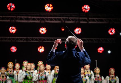 Zbliżenie na dyrygenta od tyłu, w tle widoczny fragment chóru w kostiumach łowickich