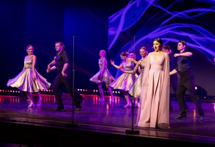 Na pierwszym planie Solistka z Chóru podczas śpiewu, za nią Artyści wykonujący choreografię taneczną