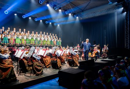 Szeroki kadr na scenę na której znajdują się Artyści Chóru w kostiumach łowickich, Artyści Orkiestry w kostiumach wilanowskich oraz Dyrygent