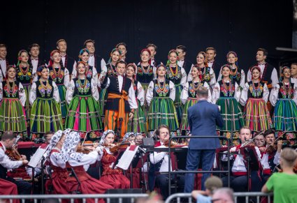 Na scenie Solista i Artyści chóru w kostiumach łowickich w trakcie śpiewu, przed nimi Dyrygent, niżej sceny Artyści Orkiestry w kostiumach wilanowskich grający na instrumentach