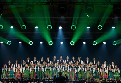 Na scenie Artyści Chóru w kostiumach łowickich, widoczny Dyrygent trzymający w górze batutę