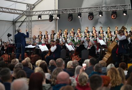 Widok z perspektywy publicznośći na chór w kostiumach łowickich, orkiestrę w kostiumach wilanowskich i dyrygującego Dyrektora zespołu