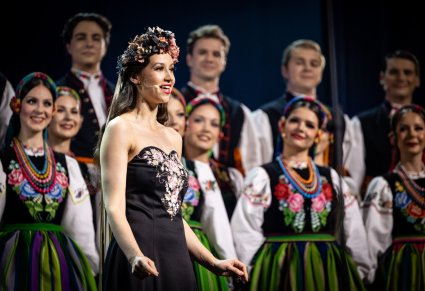 Zbliżenie na Solistkę ubraną w czarną, długą suknię i wianek na głowie, za nią widoczna część Artystów chóru w kostiumach łowickich