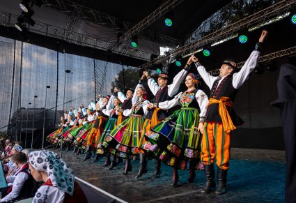Grupa Artystów w kostiumach łowickich w trakcie wykonywania choreografii tanecznej na scenie, niżej sceny widoczna część Orkiestry w kostiumach wilanowskich