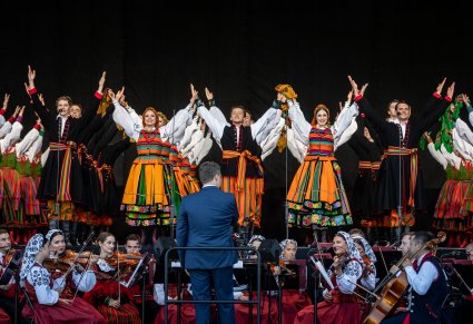 Uśmiechnięci Artyści w trakcie ukłonów w kostiumach ludowych na scenie. Na dole orkiestra wraz z Dyrygentem