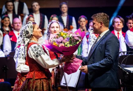Na pierwszym planie Muzyk z Orkiestry wręczająca kwiaty Dyrygentowi, w tle część Orkiestry w kostiumach wilanowskich i Chóru w kostiumach łowickich