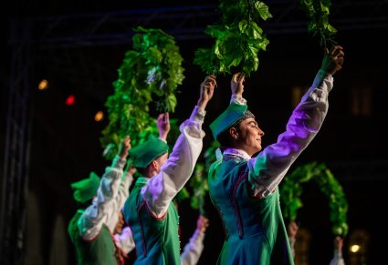 Delikatnie uśmiechnięty Artysta na scenie w kostiumie lubuskim w tle większość Artystów w tych samych kostiumach