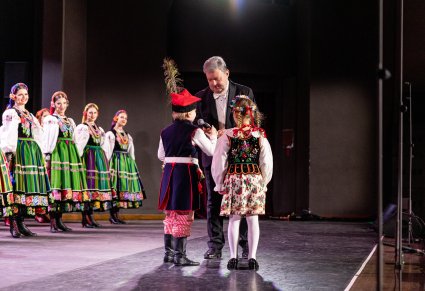 Na scenie na pierwszym planie Dyrektor zespołu trzymający mikrofon w stronę chłopca i dziewczynki. Dzieci ubrane w kostiumy krakowskie. W tle widoczna część Artystek w kostiumach łowickich