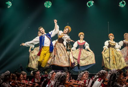 Zbliżenie ma parę Artystów w kostiumach rozbarskich a za nimi reszta Artystek w trakcie wykonywania choreografii tanecznej, widoczna część Orkiestry