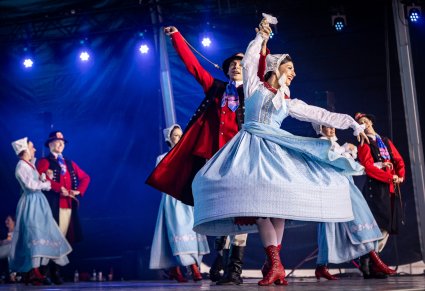 Na pierwszym planie na scenie para Artystów w kostiumach szamotulskich w trakcie wykonywania choreografii tanecznej. W tle część Artystów