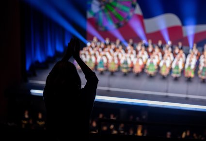 Na pierwszym planie klaszcząca osoba z publiczności a w tle na scenie zespół w kostiumach ludowych