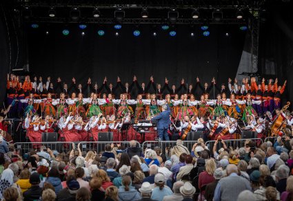 Artyści na scenie w kostiumach ludowych na dole orkiestra z Dyrektorem Jackiem Bonieckim i widoczna publiczność