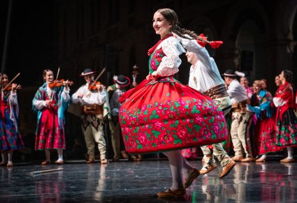 Na głównym planie Artystka w trakcie wykonywania choreografii na scenie w kostiumach podhalańskich w tle reszta Artystów