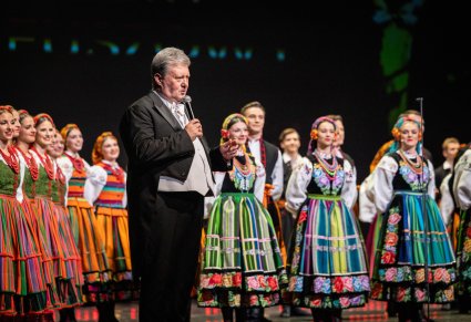 Na scenie Dyrektor zespołu w trakcie przemowy, w tle Artyści w kostiumach ludowych