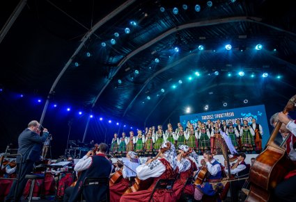 Dyrektor Jacek Boniecki dyrygujący koncert jest zwrócony w kierunku orkiestry. Za nim na scenie Artyści z chóru w kostiumach łowickich