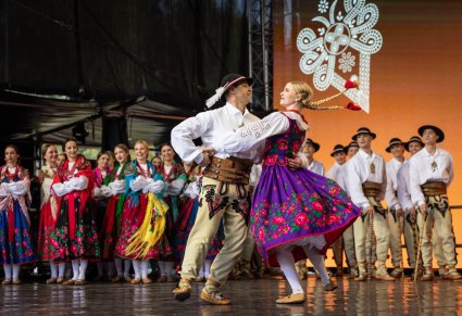 Na pierwszym planie para Artystów w kostiumach podhalańskich w trakcie wykonywania choreografii tanecznej, w drugim planie pozostała część artystów