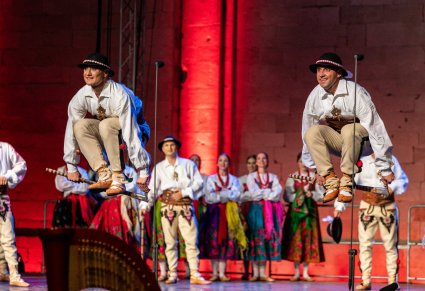Artyści na scenie w kostiumach podhalańskich na pierwszym planie dwóch Artystów wykonujących choreografię taneczną